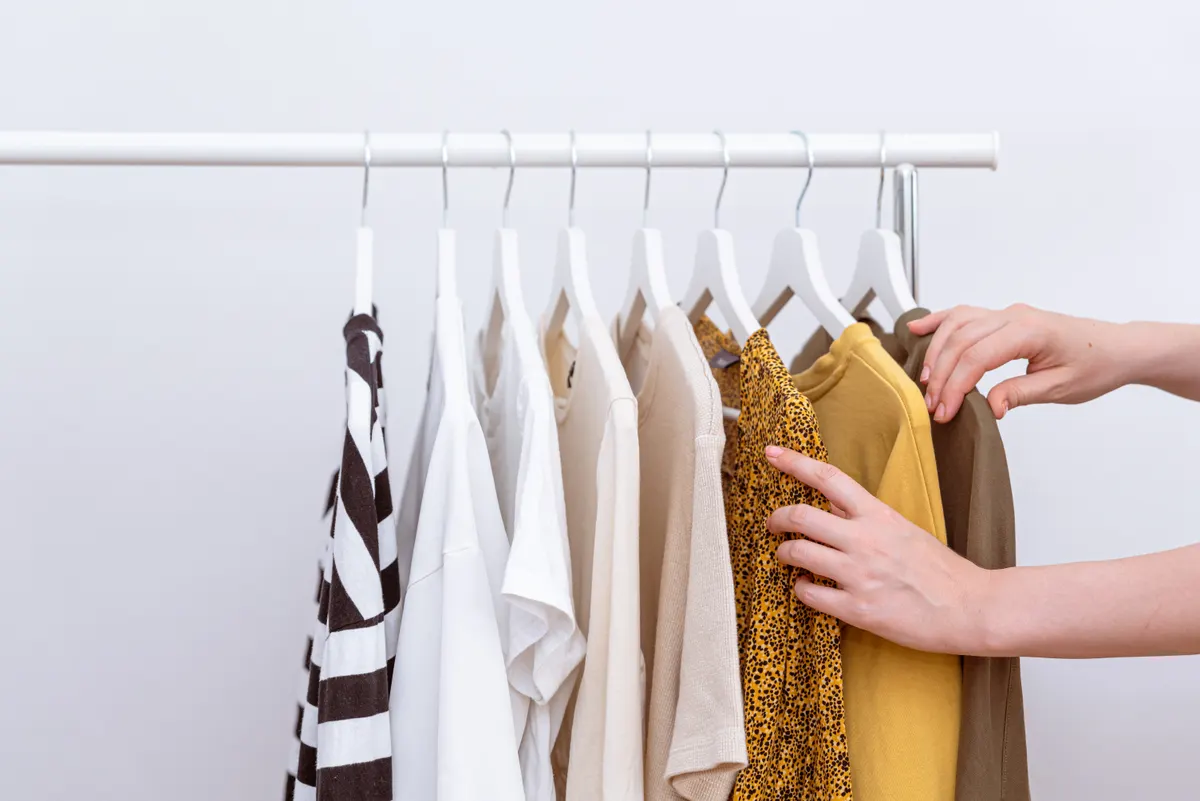 Hands selecting a mustard garment from a light clothing rail